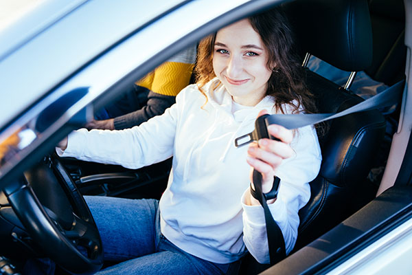 Smiling woman in a white shirt buckling her seat belt in the driver's seat of a car.